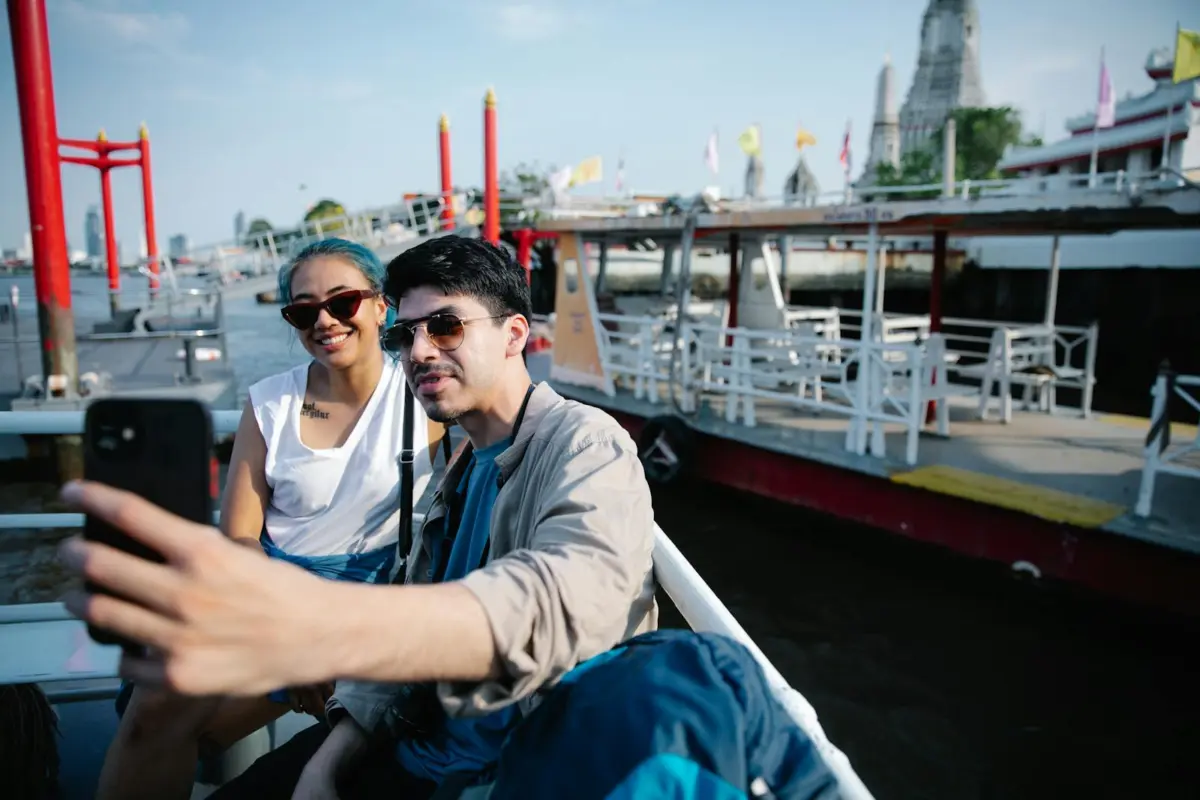 A joyful couple takes a selfie on a boat with a river and temple in the background, capturing a sunny day outdoor adventure.